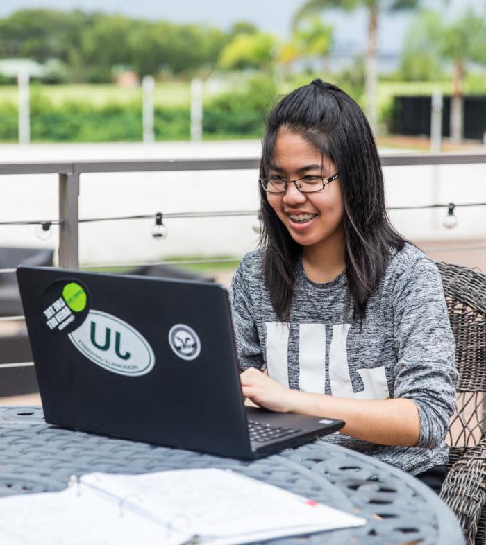 A student seating outdoors wearing a Jacksonville University t-shirt while working on their laptop. 